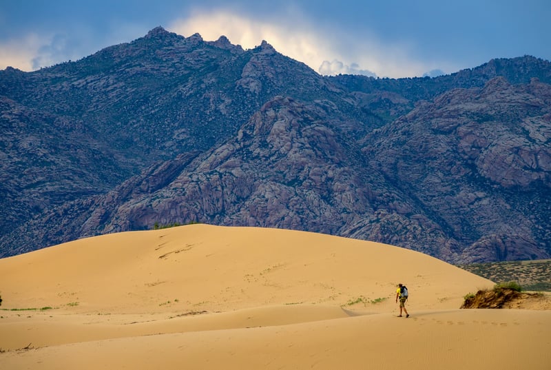 A lone hiker walking across massive sand dunes in the Gobi Desert, Mongolia, with rocky mountains in the background