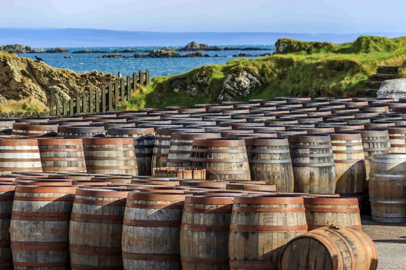 Whisky oak barrels piled high outside a distillery with the rugged Scottish coast in the background