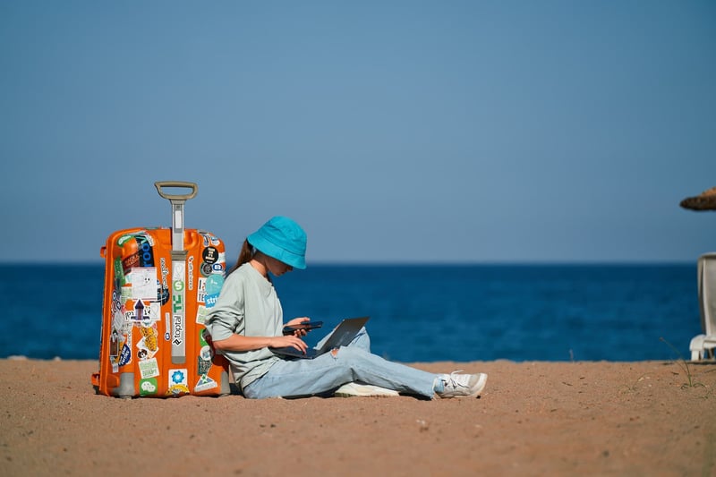 A young lady checking her phone with a laptop leaning up against her luggage on a beach
