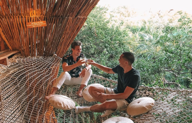 Couple of relaxed dudes clinking drinks on a massive hammock with tropical jungle in the background