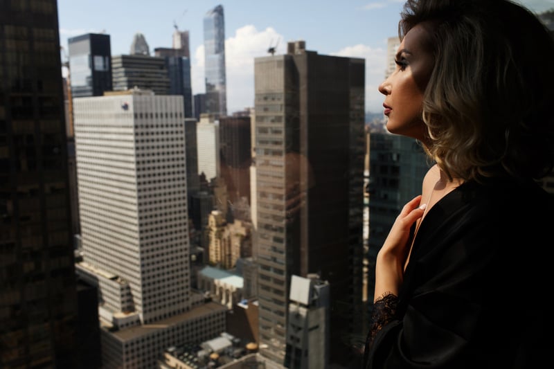 Woman in black silk robe stands with naked shoulders before a panoramic window in New York