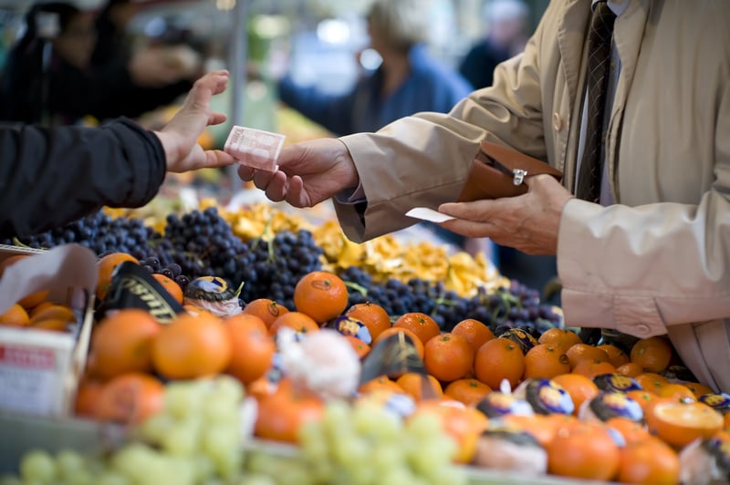 A man reaches across a row of fruit in an outdoor market to pay a vendor with a 10 Euro bill to support the local business and economy