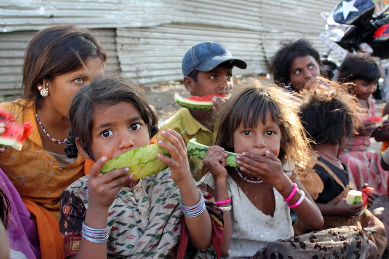 A poor girl in India eating a eatermelon along with her other family who spend their time begging on the streets